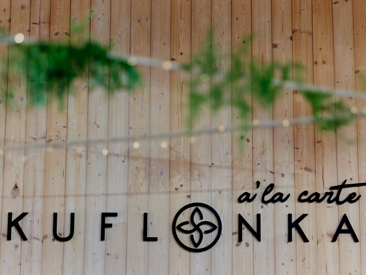 Zbliżenie na drewnianą ścianę z napisem "KUFLONKA a'la carte" w ciepłych tonach, z rozmytymi zielonymi gałązkami w przedniej części kadru